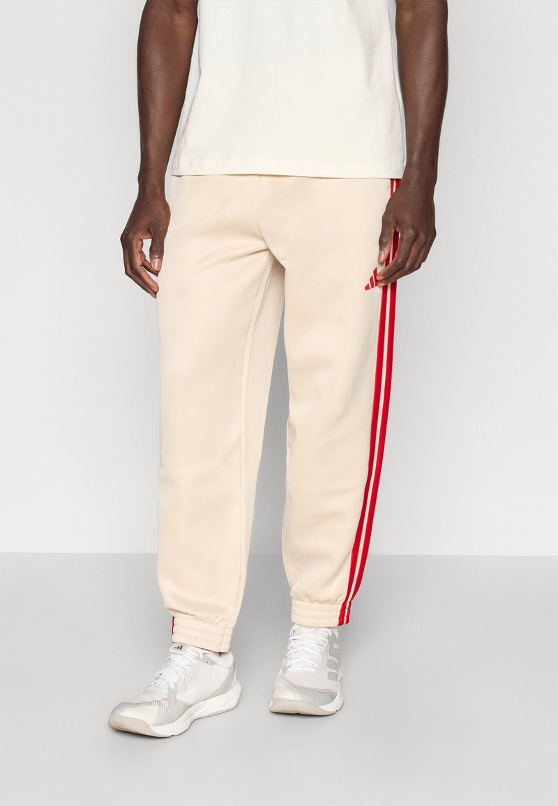 Beige Jogginghose mit zwei vertikalen roten Streifen entlang der Seiten. Mit gerippten Bündchen und einem roten Adidas-Logo auf der linken Seite.