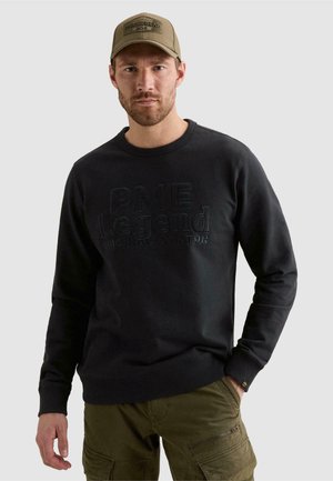 Homme portant un sweat-shirt noir PME Legend et un pantalon cargo vert olive avec casquette, debout devant un fond clair uni.