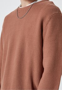 Nahaufnahme einer Person, die einen strukturierten rustfarbenen Strickpullover und eine silberne Halskette trägt, mit Fokus auf dem Oberkörper und dem Halsbereich.