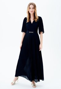 Vestido largo negro plisado con mangas cortas acampanadas y escote en V, que presenta un detalle de cinturón metálico en la cintura. Textura de tela suave.