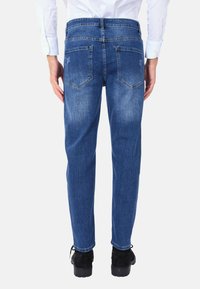 Jeans in denim blu visto da dietro, con un taglio dritto, due tasche posteriori e una leggera scoloritura sulle cosce. Scarpe nere visibili.