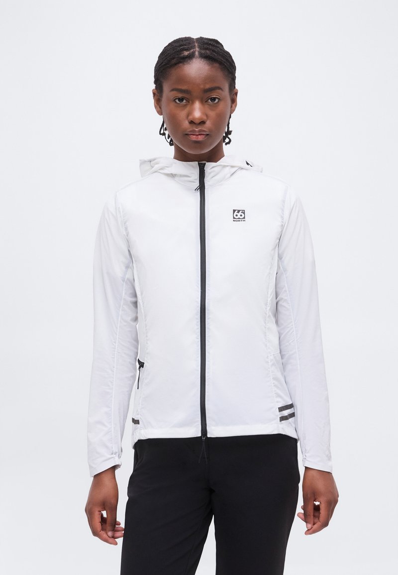 Femme avec des tresses portant une veste blanche à capuche avec une fermeture éclair noire et un logo, associée à un pantalon noir, debout devant un fond uni.