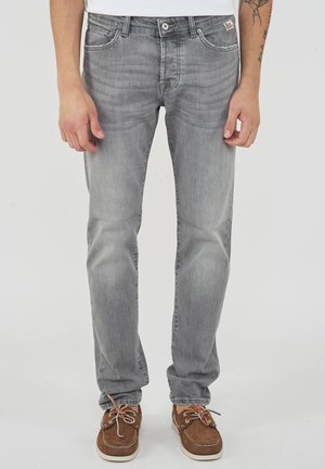 Uomo che indossa jeans slim fit grigio chiaro con scarpe da barca marroni, in piedi davanti a uno sfondo bianco semplice, con le mani rilassate ai lati.