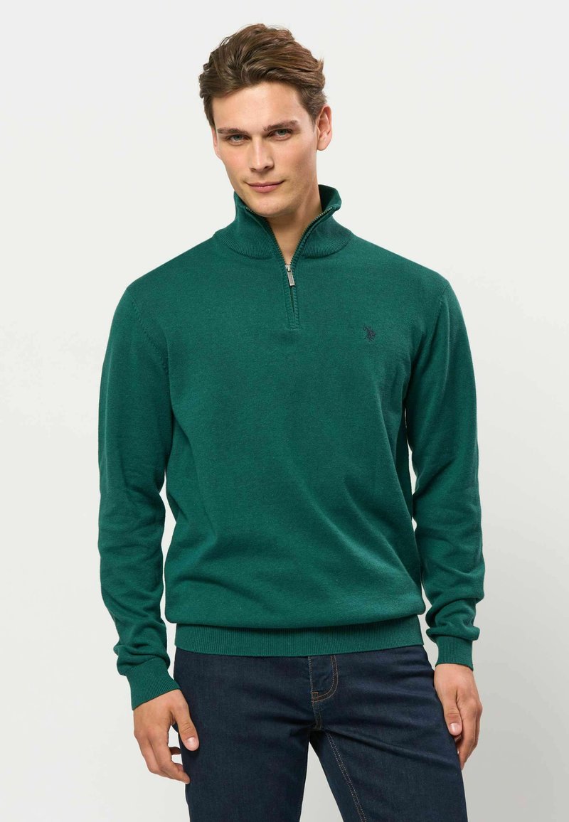 Homme aux cheveux bruns courts portant un pull vert zippé et un jean bleu foncé, debout devant un fond clair uni.