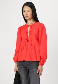 Blusa di cotone rossa con maniche a palloncino e dettagli a chiave sul davanti, con lacci. L'orlo è arricciato, indossata sopra jeans grigi scuri.