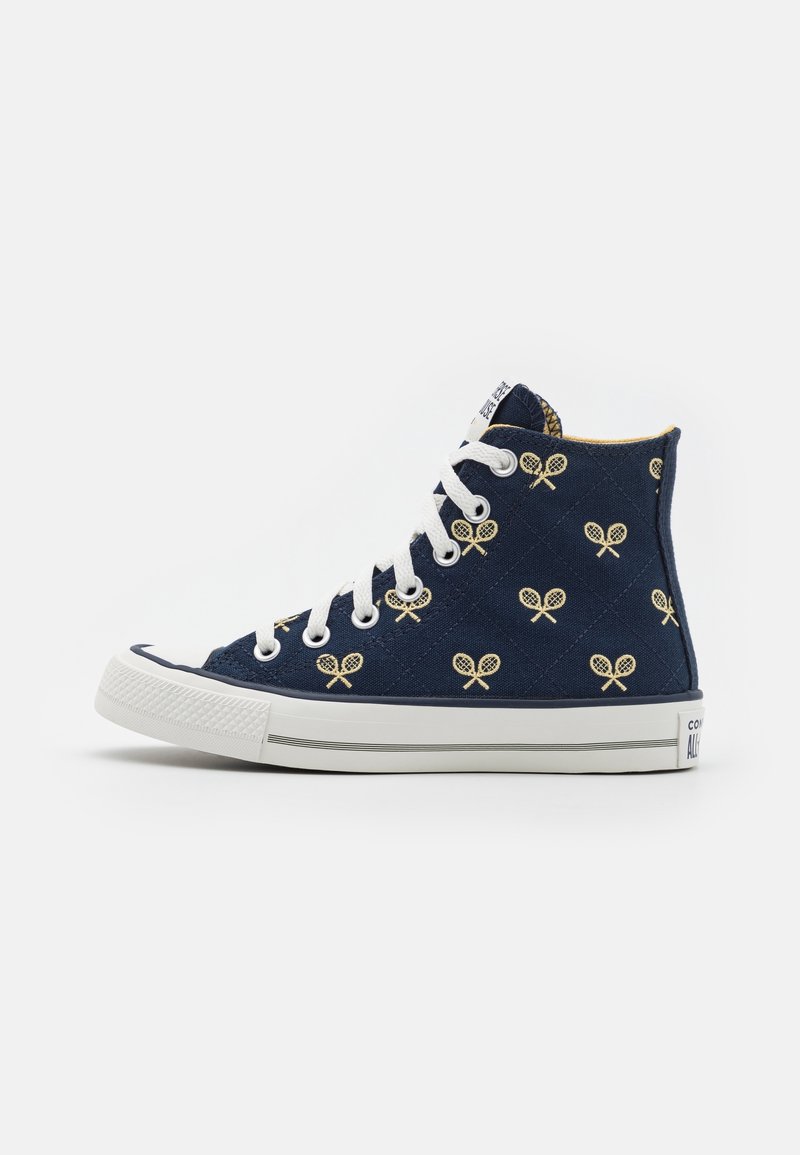 Baskets montantes en toile marine avec des lacets blancs, présentant un motif matelassé et des accents de raquette de tennis jaunes. Capuchon en caoutchouc à l'avant et semelle.