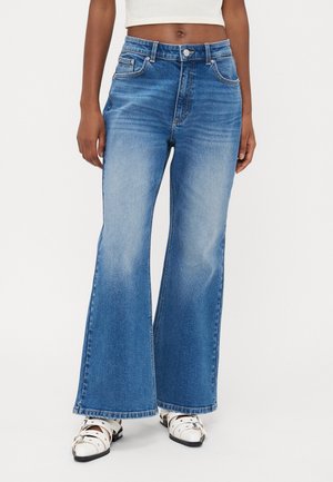 Femme portant un jean bleu taille haute à jambes larges avec un devant délavé et des chaussures blanches pointues à brides, sur un fond blanc.