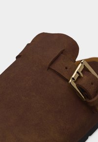 Καφέ δερμάτινο παπούτσι σουέτ με χρυσή μεταλλική αγκράφα στο λουράκι, που αναδεικνύει την υφή και τη λεπτομέρεια του κλεισίματος.