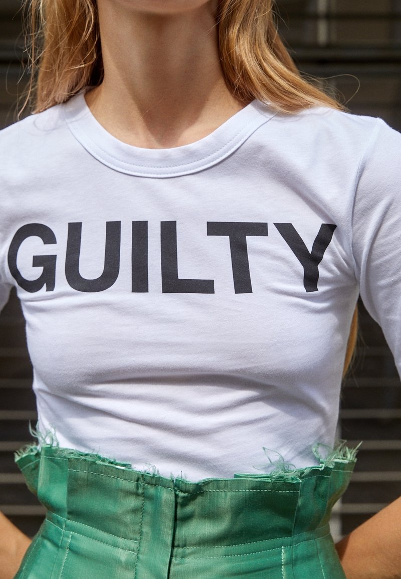 Camiseta de algodón blanca con un estampado negro en negrita que dice "CULPABLE". Combinada con una falda de tela verde texturizada de cintura alta con bordes deshilachados.