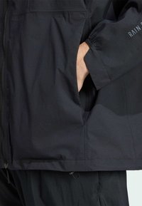 Jachetă de ploaie neagră cu țesătură texturată, închidere cu fermoar, manșete elastice și buzunare laterale. Prezintă un brand discret pe mânecă.
