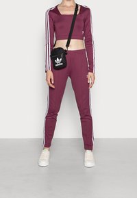 Bordový sportovní set s dlouhým rukávem a crop topem a kalhotami s bílými pruhy. Modelka nese malou černou kabelku přes rameno s logem.