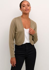 Cárdigan corto de color verde oliva claro, hecho de un material de punto texturizado, con un frente redondeado y mangas largas, que se combina con una camiseta blanca y pantalones negros.