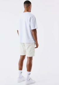 Camiseta blanca de manga corta, corte holgado, combinada con pantalones cortos cargo beige claros. Los accesorios incluyen zapatillas blancas y calcetines con detalles en negro.