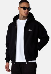 Sweat à capuche noir zippé avec une grande capuche, logo brodé blanc sur la poitrine gauche et poignets côtelés. Porté par-dessus une chemise blanche.