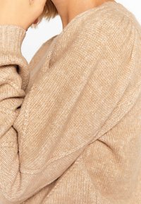 Beige stickad tröja med en mjuk textur, ribbade ärmslut och en avslappnad passform. Synlig sömnadsdetalj ger subtilt designintresse.