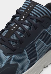 Κοντινό πλάνο μπλε και μαύρου αθλητικού παπουτσιού με μαύρα κορδόνια και μαύρη θηλιά στην γλώσσα με λευκό κείμενο που γράφει «TRAIL UNDER».