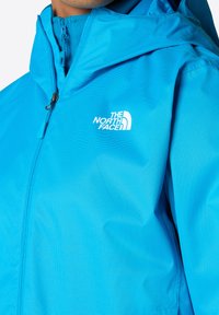 Světle modrá bunda vyrobená z syntetického materiálu s texturovaným povrchem, vybavená zapínáním na zip a kapucí. Logo je výrazně umístěno.