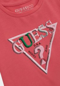 Koraļļu T-krekls, kas izgatavots no organiskās kokvilnas, ar trīsstūrveida izšūtu plāksteri, kurā ir sekvīnu "GUESS" logo, cipari un jautājuma zīmes dizains.