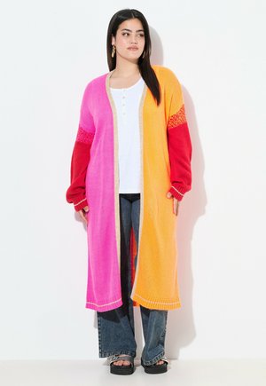 Donna che indossa un cardigan lungo colorato nei toni del rosa, arancione e rosso, una maglia henley bianca, jeans svasati e sandali con piattaforma, in piedi contro un muro bianco.
