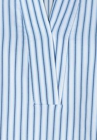 Helle blaue Stoff mit vertikalen navyblauen Streifen. Verfügt über ein V-Ausschnitt-Design und ein dezentes Taschen-Detail, das eine glatte und weiche Textur zur Schau stellt.