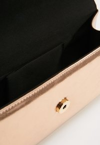 Clutch metálica color oro rosa abierto con interior de tela negra y un cierre magnético redondo dorado sobre una superficie blanca.