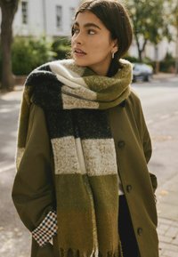 Écharpe à motif à carreaux vert, noir et crème, en laine douce, drapée autour du cou, avec des franges aux extrémités.