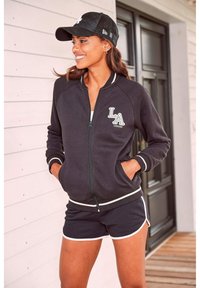 Čierna zipová bunda so bielymi pruhmi, zdobená výšivkou "LA", v kombinácii s čiernymi šortkami lemovanými bielou farbou. Nosí sa s čiernym klobúkom.