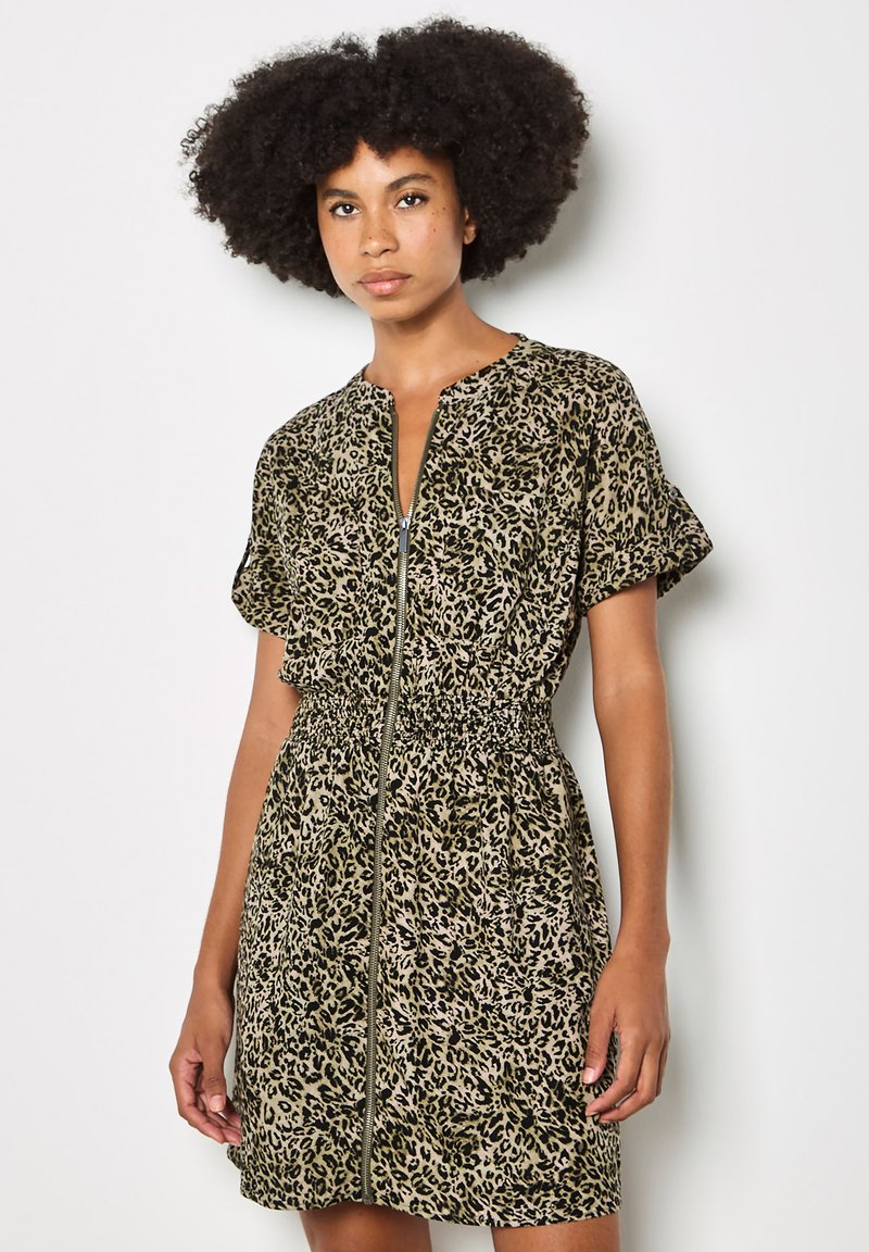 Femme avec une coiffure afro naturelle portant une robe léopard à manches courtes avec fermeture éclair sur le devant et taille froncée, debout devant un fond blanc.
