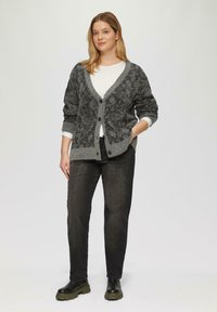 Grijze gebreide cardigan met een V-hals, knoopsluiting en een patroontextuur, gecombineerd met een witte top, zwarte jeans en stevige zwarte laarzen.
