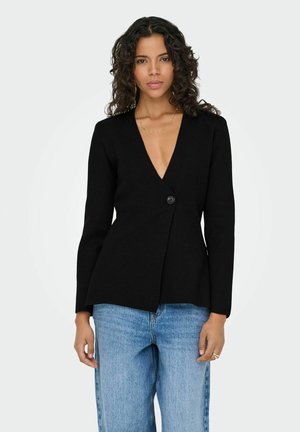 Femme aux cheveux bouclés portant une veste noire à un seul bouton et un jean bleu clair, debout devant un fond clair uni.