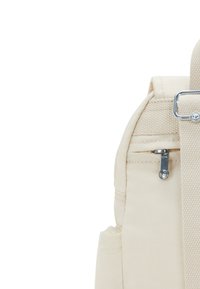 Mochila de tela beige con un bolsillo de cremallera lateral, superficie texturada y una correa ancha ajustable. Presenta herrajes plateados y detalles de costura.