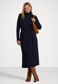 Donna che indossa un abito midi a dolcevita blu navy, stivali marroni sopra il ginocchio, orecchini a cerchio e porta una borsa a tracolla marrone.