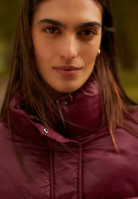 Bordeaux puffer jacket met een hoge kraag, voorzien van een ritssluiting en drukknoopdetails, gemaakt van glanzend, gewatteerd materiaal.