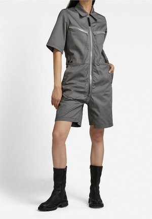 Personne portant une combinaison grise à manches courtes avec fermeture éclair devant et poches zippées, ainsi que des bottes en cuir noires mi-mollet, debout sur un fond blanc.