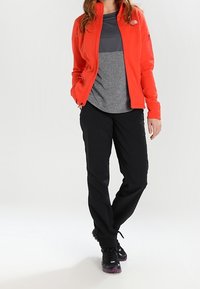 Chaqueta roja de cremallera, camiseta gris de manga corta, pantalones negros y zapatillas deportivas negras con acentos morados, que lleva puesta una persona de pie contra un fondo claro.