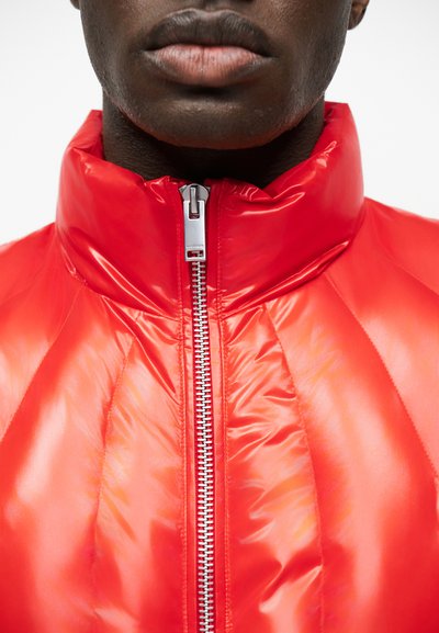 Κοντινό πλάνο ενός ατόμου που φοράει λαμπερό κόκκινο μπουφάν με επένδυση και ασημένιο εμπρόσθιο φερμουάρ, εμφανιζόμενο από το πηγούνι έως το στήθος.