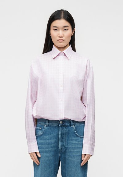 Femme portant une chemise rose clair à boutons avec une poche poitrine en forme de cœur, associée à un jean bleu, debout devant un fond uni.