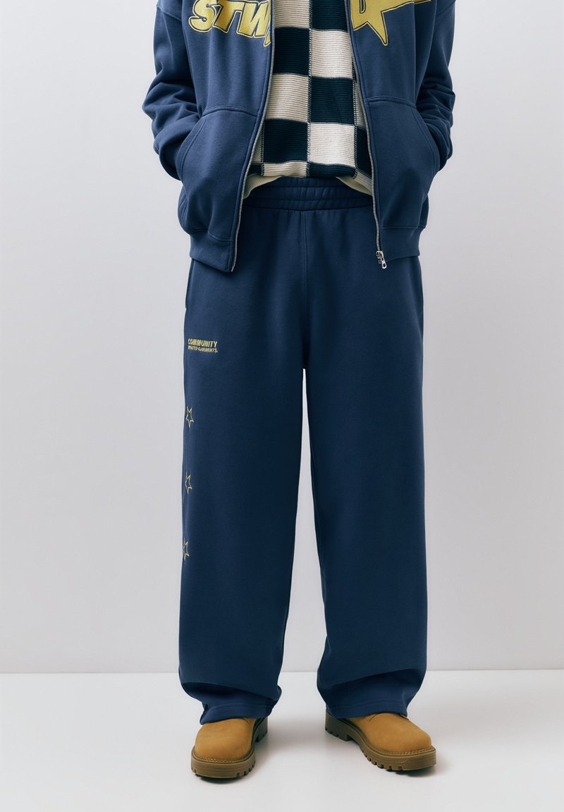 Persoon die marineblauwe joggingbroek met gele sterdetails, zwart-wit geruit vest, marineblauwe capuchontrui met rits en tankleurige laarzen draagt, staand.