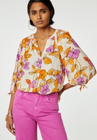 Bloemrijke blouse met oranje en paarse patronen op een crèmekleurige achtergrond, met een V-hals en driekwart pofmouwen; gecombineerd met roze jeans.