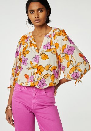 Blumige Bluse mit orangefarbenen und lila Mustern auf einem cremefarbenen Hintergrund, mit einem V-Ausschnitt und dreiviertellangen Puffärmeln; kombiniert mit rosa Jeans.