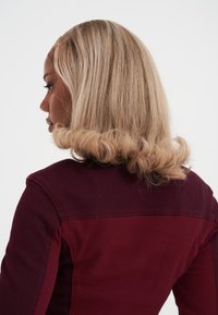 Kobieta z włosami do ramion, blondynką, z lokami na końcach, w bordowej kurtce, stojąca w lewo na białym tle.