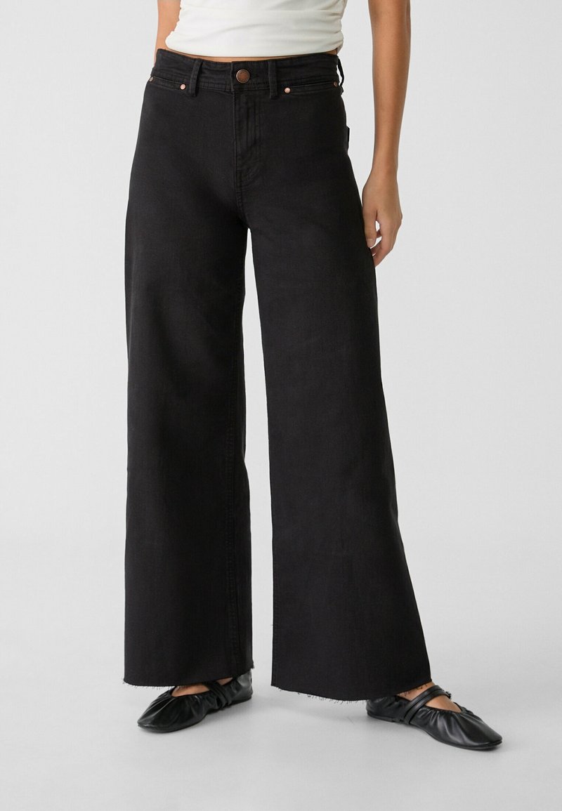 Personne portant un jean noir taille haute à jambes larges et des ballerines noires à fines lanières, debout devant un fond clair uni.