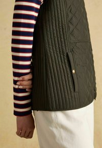 Prešívaná zelená vesta s vertikálnymi a diamantovými vzormi, s bočnými vreckami a detailom gombíka, nosená cez modro-červenú prúžkovanú košeľu.