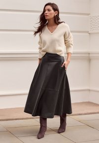 Beiger Rippstrickpullover mit V-Ausschnitt kombiniert mit einem schwarzen Lederrock in A-Linie, der seitliche Taschen und eine glatte Textur aufweist. Dazu werden lila Wildlederstiefel getragen.