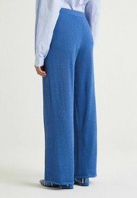 Pantalon large bleu en tissu doux et texturé avec une taille haute. La vue de dos révèle un design épuré sans ornements visibles.