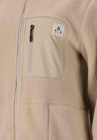 Beige fleecejakke med en lynlåslomme på brystet og et stofdetalje. Inkluderer logo-patch og tekstureret materiale for varme.