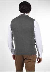 Grauer Strickpullover ohne Ärmel mit einem gerippten Kragen und Saum, getragen über einem weißen Langarmshirt, kombiniert mit beigen Hosen.
