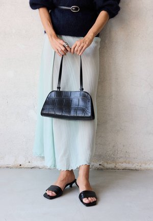 Femme portant une jupe bleu clair et un pull foncé tient un sac à main noir texturé, portant des sandales noires à talons sur un sol en béton.