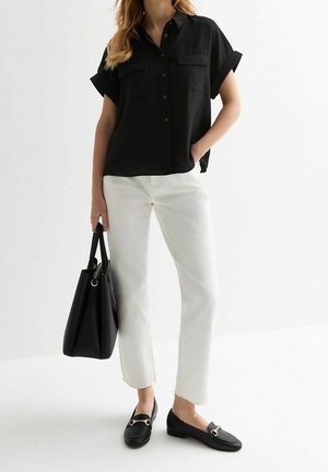 Kvinde iført en sort kortærmet skjorte med knapper, hvide cropped bukser, sorte loafers og bærende på en stor sort tote taske.