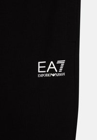 Crne trenirke od mekog materijala, s bijelim logotipom "EA7" i "EMPORIO ARMANI" otisnutim na donjem dijelu nogavice. Podesiv remen.